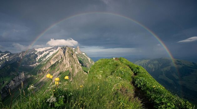 A vivid rainbow spans a dark, stormy sky, arching over the jagged peaks of the Alpstein mountains. Switzerland. Photo by Pascal Debrunner