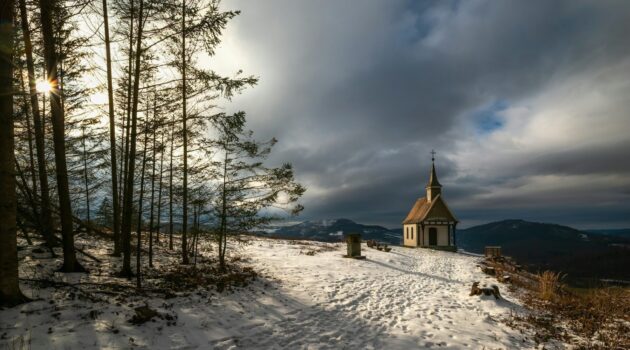 Small chapel on a snowy hill under dramatic clouds. Photo by Evgeni Tcherkasski