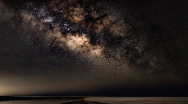 The Milky Way stretches above a quiet coastal inlet as soft reflections fade into the night. Photo by Dmytro Koplyk