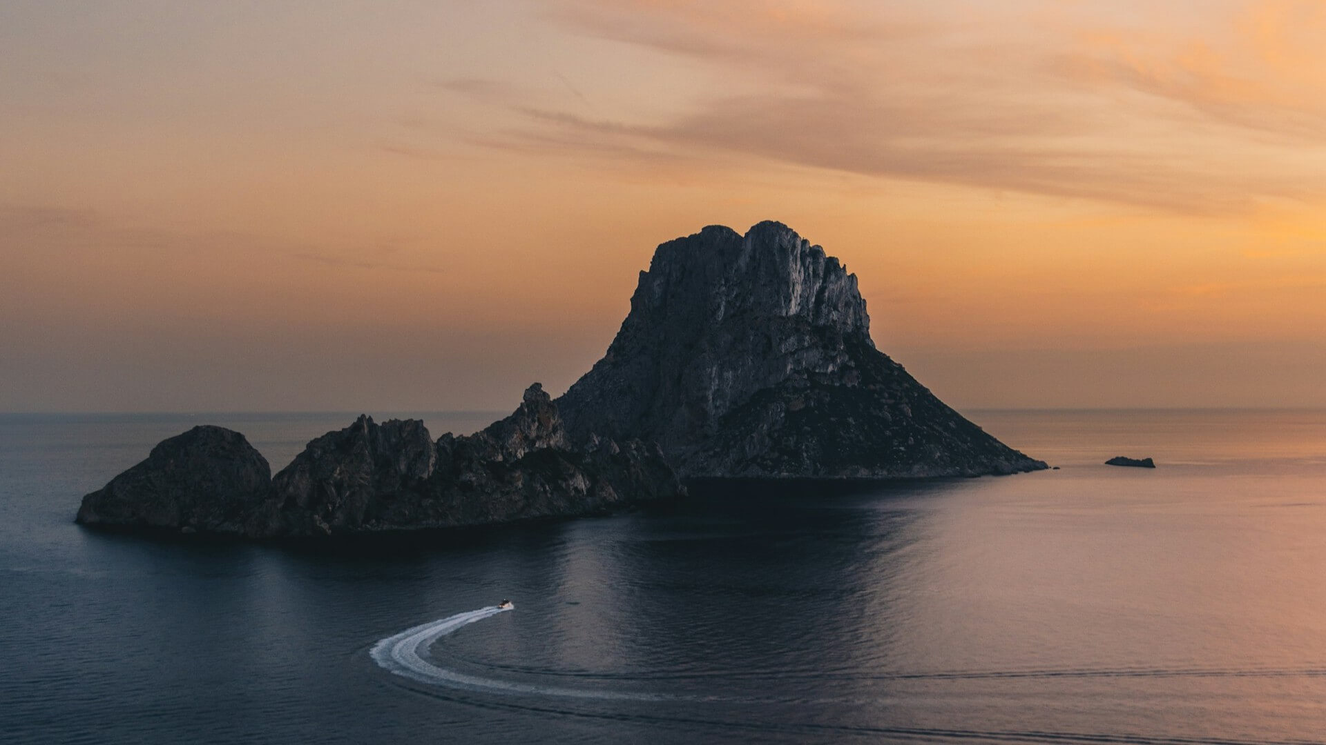 A speedboat carves a graceful arc across the calm waters below Es Vedrà as the Mediterranean glows beneath a vibrant Ibiza sunset. Isla de es Vedrà, Spain. Photo by Damian Kamp